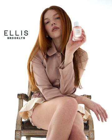 Woman with red hair wearing a tan jacket and cream skort sits in a wooden chair and holds VANILLA RICE Eau de Parfum 50ml against a white background with a black Ellis Brooklyn logo. 