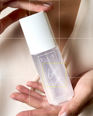 Closeup of model wearing white a white shirt and holding COCOA BLANCO Perfume Mist between her hands against her chest. The photo is overlaid with white and yellow camera grid lines. 