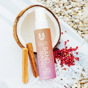 Brume parfumée pour cheveux et corps Sand 50ml s'inspirant de la noix de coco, aux côtés du bois de santal, des fleurs blanches et du poivre rose sur une serviette rayée bleu et blanc, avec des cailloux en arrière-plan.