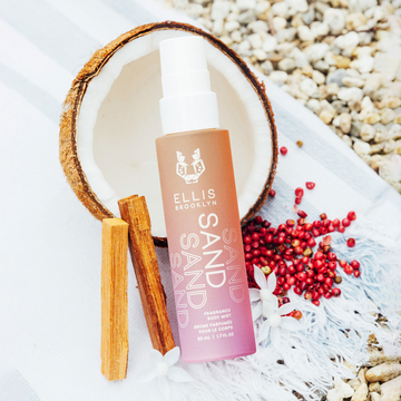 Brume parfumée pour cheveux et corps Sand 50ml s'inspirant de la noix de coco, aux côtés du bois de santal, des fleurs blanches et du poivre rose sur une serviette rayée bleu et blanc, avec des cailloux en arrière-plan.