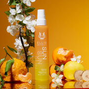 Brume parfumée pour cheveux et corps Sun 100 ml sur un miroir à côté d'oranges, de citrons, de bois de cèdre, de fleurs blanches, de feuilles vertes et de gousses de vanille, sur un fond dégradé orange et jaune.