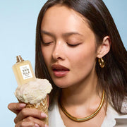 Une femme aux cheveux noirs, vêtue d'une chemise blanche et portant des créoles ainsi qu'un collier en or, tient un cornet de glace à la vanille surmonté du flacon d'eau de parfum VANILLA SALT (7,5 ml, sans bouchon), le tout sur fond bleu.
