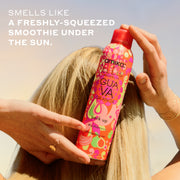 Woman with blonde hair and gold ring and bracelets holds Amika x Ellis Brooklyn GUAVA GRANITA Perk Up Dry Shampoo against the back of her head against blue and yellow sky with white text that reads 
