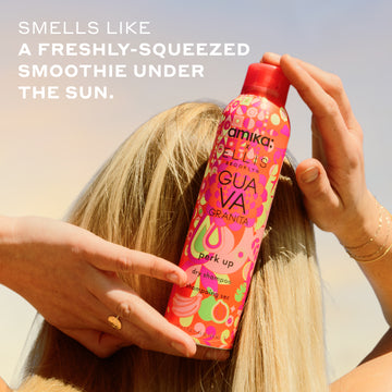 Woman with blonde hair and gold ring and bracelets holds Amika x Ellis Brooklyn GUAVA GRANITA Perk Up Dry Shampoo against the back of her head against blue and yellow sky with white text that reads 