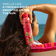 Woman with brown hair wearing a white tank top sits on white, pink, and orange beach chair and holds amika x Ellis Brooklyn GUAVA GRANITA Perk Up Dry Shampoo next to her head against background of blue ocean and sky with white text that reads 