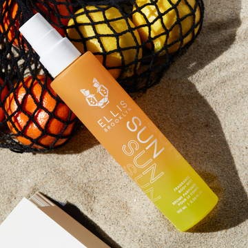 Brume parfumée pour cheveux et corps Sun 100ml couleur sable, appuyée sur un sac en filet noir rempli d'oranges et de citrons, près du coin d'un livre ouvert.