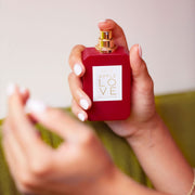 Model with light pink nail polish holding and spraying Apple Love 50ml Eau De Parfum in one hand, against green couch and white wall.