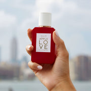 Model with light pink nail polish holding Apple Love 50ml Eau De Pafum with city in background.