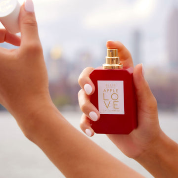 Model with light pink nail polish holding and spraying Apple Love 50ml Eau De Parfum in one hand while holding cap in the other, with city in background.