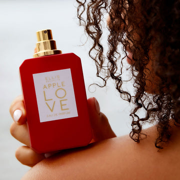 Model with brown curly hair and light pink nail polish holding Apple Love 50ml  Eau De Parfum with cap removed on shoulder, against white background.