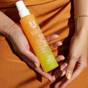 Modèle avec du vernis à ongles jaune portant des bracelets en or et une robe orange tenant un flacon de 100 ml de brume parfumée pour cheveux et corps Sun dans une main sur les genoux.