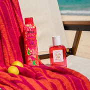 Amika x Ellis Brooklyn GUAVA GRANITA Perk Up Dry Shampoo and GUAVA GRANITA 50ml Eau de Parfum sit on white and brown beach chair next to orange and pink beach towel and lemons against background of blue ocean and tan sand