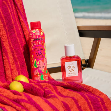 Amika x Ellis Brooklyn GUAVA GRANITA Perk Up Dry Shampoo and GUAVA GRANITA 50ml Eau de Parfum sit on white and brown beach chair next to orange and pink beach towel and lemons against background of blue ocean and tan sand