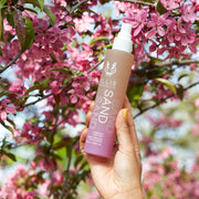 Model with yellow nail polish wearing gold ring holding Sand Hair and Body Fragrance Mist 100ml in front of tree branches with pink flowers, on blue background.