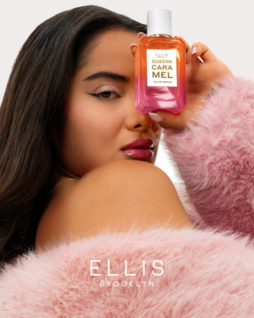 Model with black hair and French manicure wearing pink fur jacket and gold necklace holding Queens Caramel 50ml Eau De Parfum in front of face, on white background.