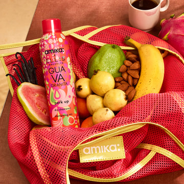 Amika x Ellis Brooklyn GUAVA GRANITA Perk Up Dry Shampoo in pink and yellow mesh amika bag with guavas, vanilla beans, almonds, a banana, and oranges next to a dragonfruit and white mug with a pink lipstick stain filled with coffee on a brown table