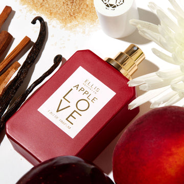 Apple Love 50ml Eau De Parfum with cap removed next to white flower, apple, plum, sandal wood, vanilla beans, and sugar on white background.