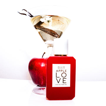 Apple Love 50ml Eau De Parfum on white table in front of apple and martini glass holding white flower, vanilla bean, and water, against white background.