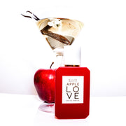 Apple Love 50ml Eau de Parfum sur une table blanche devant une pomme et un verre à martini tenant une fleur blanche, une gousse de vanille et de l'eau, sur fond blanc.