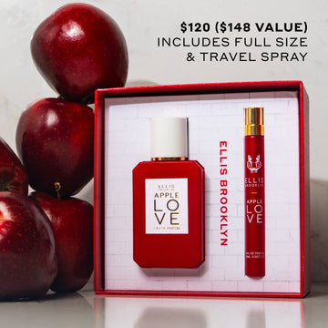 Apple Love Eau De Parfum Gift Set Box showing Apple Love 50ml Eau De Parfum and Apple Love 10ml Eau De Parfum Travel Spray on white table next to stack of red apples on white background with black text that reads 