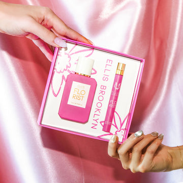 Model with nude nail polish holding top corner of Florist Eau De Parfum Gift Set Box showing Florist 50ml Eau De Parfum and Florist 10ml Eau De Parfum Travel Spray and model with grey iridescent nail polish holding bottom corner of same box, against pink satin background.