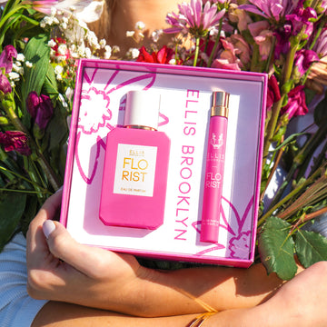 Model with blonde hair and nude nail polish, wearing white long sleeve shirt and gold bracelets holding Florist Eau De Parfum Gift Set Box showing Florist 50ml Eau De Parfum and Florist 10ml Eau De Parfum Travel Spray and bouquets of red, pink, and white flowers in both arms against chest. | english_image