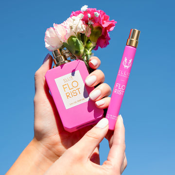 Model with nude nail polish holding Florist 50ml Eau De Parfum with cap removed with pink and white flowers in one hand and Florist 10ml Eau De Parfum Travel Spay in other hand, on blue background.