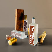 Huile parfumée Vanilla Santo (15 ml, bouchon retiré) à côté de bois de palo santo, huile parfumée Blond Musk (15 ml, bouchon retiré), ambre blond, huile parfumée Milk Wood (15 ml) et un morceau de bois sur fond brun.