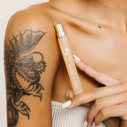 Modèle portant un haut beige sans bretelles avec un tatouage de dragon et du vernis à ongles blanc tenant le spray voyage Vanilla Milk 10ml Eau De Parfum sous la clavicule sur fond blanc.