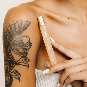Modèle portant un haut beige sans bretelles avec un tatouage de dragon et du vernis à ongles blanc tenant le spray voyage Vanilla Milk 10ml Eau De Parfum sous la clavicule sur fond blanc.