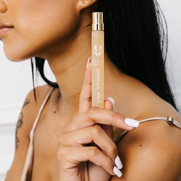 Un modèle aux cheveux noirs, avec du vernis blanc et un tatouage sur le bras, porte un débardeur en satin beige et tient le spray voyage Vanilla Milk 10 ml Eau De Parfum près de l'épaule, sur un fond blanc