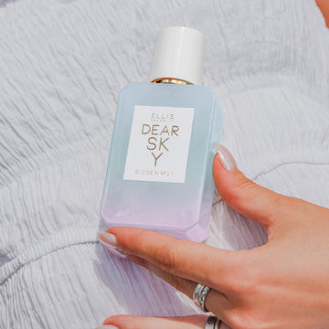 Un mannequin avec du vernis à ongles nude, portant des bagues argentées et une robe blanche, tient le parfum Dear Sky 50ml contre son ventre.