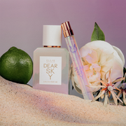 Chers parfum Dear Sky 50 ml Eau de Parfum et vaporisateur de voyage Dear Sky 10 ml Eau de Parfum dans le sable à côté de la bergamote et des fleurs, sur un fond violet.