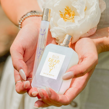 Model with nude nail polish, wearing silver rings, gold and silver bracelets, and white dress, holding Dear Sky 10ml Eau De Parfum Travel Spray, Dear Sky 50ml Eau De Parfum with cap removed, and white flower in both hands.