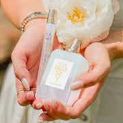 Modèle avec du vernis à ongles nu, portant des bagues en argent, des bracelets en or et en argent, et une robe blanche, tenant Dear Sky 10ml Eau De Parfum Spray de Voyage, Dear Sky 50ml Eau De Parfum avec le bouchon retiré, et une fleur blanche dans les deux mains.