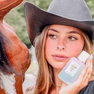 Un modèle aux cheveux bruns et au vernis à ongles blanc, portant une chemise noire, un chapeau de cowboy marron, un collier en or et une bague en argent, tient le parfum Dear Sky 50ml près du visage, tout en se tenant à côté d'un cheval marron et blanc aux cheveux noirs, avec des arbres et une route en arrière-plan.