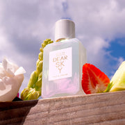 Dear Sky 50ml Eau De Parfum on piece of wood next to strawberry, melon, and flowers, with sky and clouds in the background.