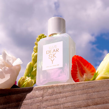 Dear Sky 50ml Eau De Parfum on piece of wood next to strawberry, melon, and flowers, with sky and clouds in the background.