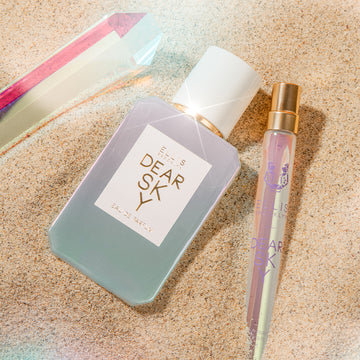Dear Sky 50ml Eau de Parfum et Dear Sky 10ml Eau de Parfum Travel Spray légèrement enterrés dans le sable à côté d'un cristal.
