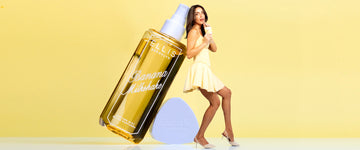Model with brown hair in yellow dress and yellow heels drinks a yellow milkshake in a glass cup with a straw, faces to the side and holds up with her back a life size bottle of BANANA MILKSHAKE Perfume Mist without a cap next to it's cap, on yellow background.