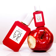 Apple Love 7.5ml Eau De Parfum Mini Dabber on apple with bite taken out of it next to Apple Love 50ml Eau De Parfum on white table, on white background.