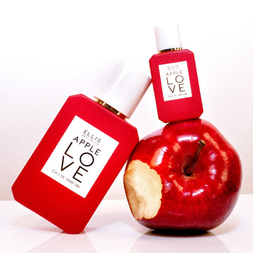 Apple Love 7.5ml Eau De Parfum Mini Dabber on apple with bite taken out of it next to Apple Love 50ml Eau De Parfum on white table, on white background.