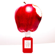 Apple with bite taken out of it on top of Apple Love 7.5ml Eau De Parfum Mini Dabber on white table, on white background.