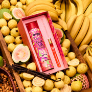 Le coffret cadeau Amika x Ellis Brooklyn GUAVA GRANITA Shampooing Sec Perk Up et GUAVA GRANITA Eau de Parfum 10 ml est posé dans une caisse en bois brun remplie de bananes, de goyaves entières et tranchées, d’un bouquet de gousses de vanille et d’amandes dans un panier brun.