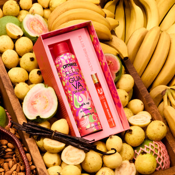 Le coffret cadeau Amika x Ellis Brooklyn GUAVA GRANITA Shampooing Sec Perk Up et GUAVA GRANITA Eau de Parfum 10 ml est posé dans une caisse en bois brun remplie de bananes, de goyaves entières et tranchées, d’un bouquet de gousses de vanille et d’amandes dans un panier brun.