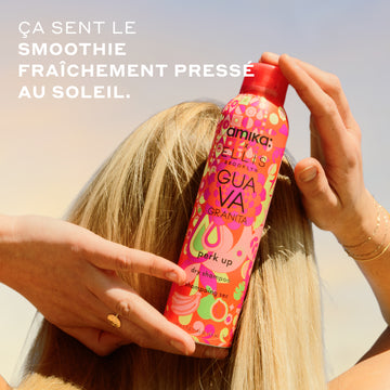 Femme aux cheveux blonds avec une bague en or et des bracelets tient le shampooing sec Amika x Ellis Brooklyn GUAVA GRANITA Perk Up contre l'arrière de sa tête, devant un ciel bleu et jaune avec un texte blanc qui dit « Ça sent le smoothie fraîchement pressé au soleil »