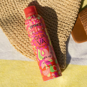 Le shampoing sec Amika x Ellis Brooklyn GUAVA GRANITA est posé sur une serviette de plage à rayures jaunes et blanches et un chapeau tressé beige
