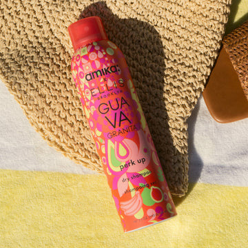 Le shampoing sec Amika x Ellis Brooklyn GUAVA GRANITA est posé sur une serviette de plage à rayures jaunes et blanches et un chapeau tressé beige