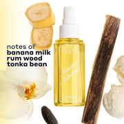 BANANA MILKSHAKE Perfume Mist 240ml without a cap surrounded by 2 slices of banana, a piece of rum wood, 2 scoops of vanilla ice cream, a tonka bean, and a flower on a white and yellow ombré background with black text that reads 