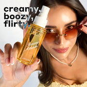 Model with brown hair wearing brown sunglasses, a yellow dress, and a pearl necklace looks up at the camera while holding BANANA MILKSHAKE 90ml perfume mist without a cap on a blue and white ombre background with black, white, and blue text that reads 